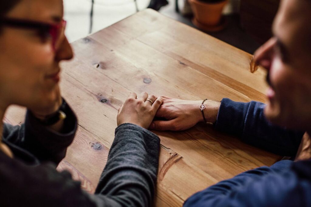 Two people are visible holding hands on a wooden table while talking to each other.